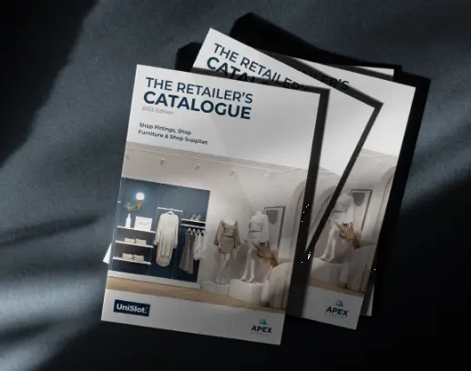 Two copies of The Retailers Catalogue 2023 edition rest on a dark surface. The cover features a retail display with mannequins, clothing racks, and shelves. Logos of Unislot and Apex stand out. Sunlight delicately casts shadows across the captivating scene.