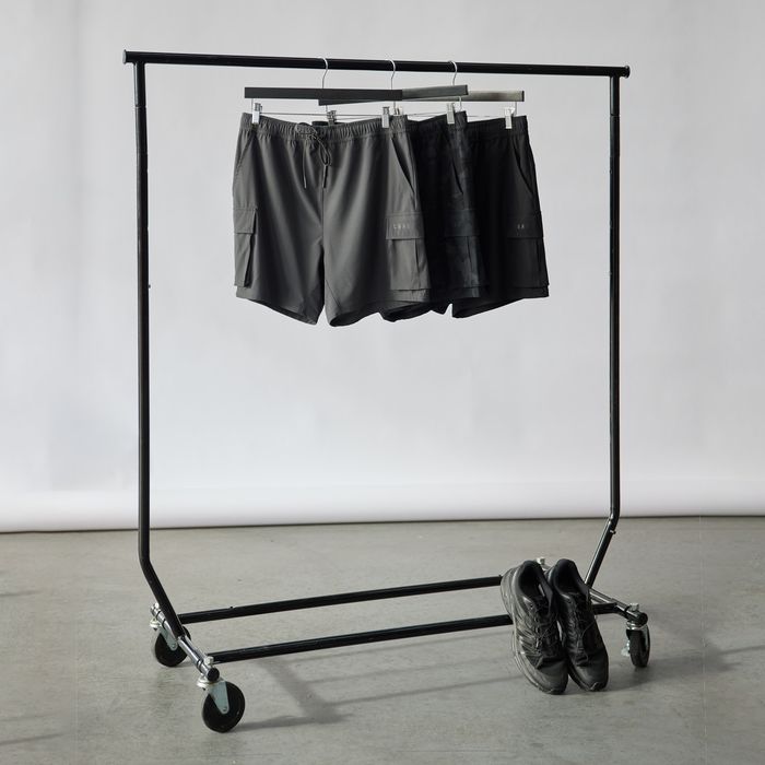 A Single Rail Travellers Rack Black holds three pairs of dark shorts on hangers. Below the folding rack, a pair of black shoes rests against a plain light-colored wall and floor.