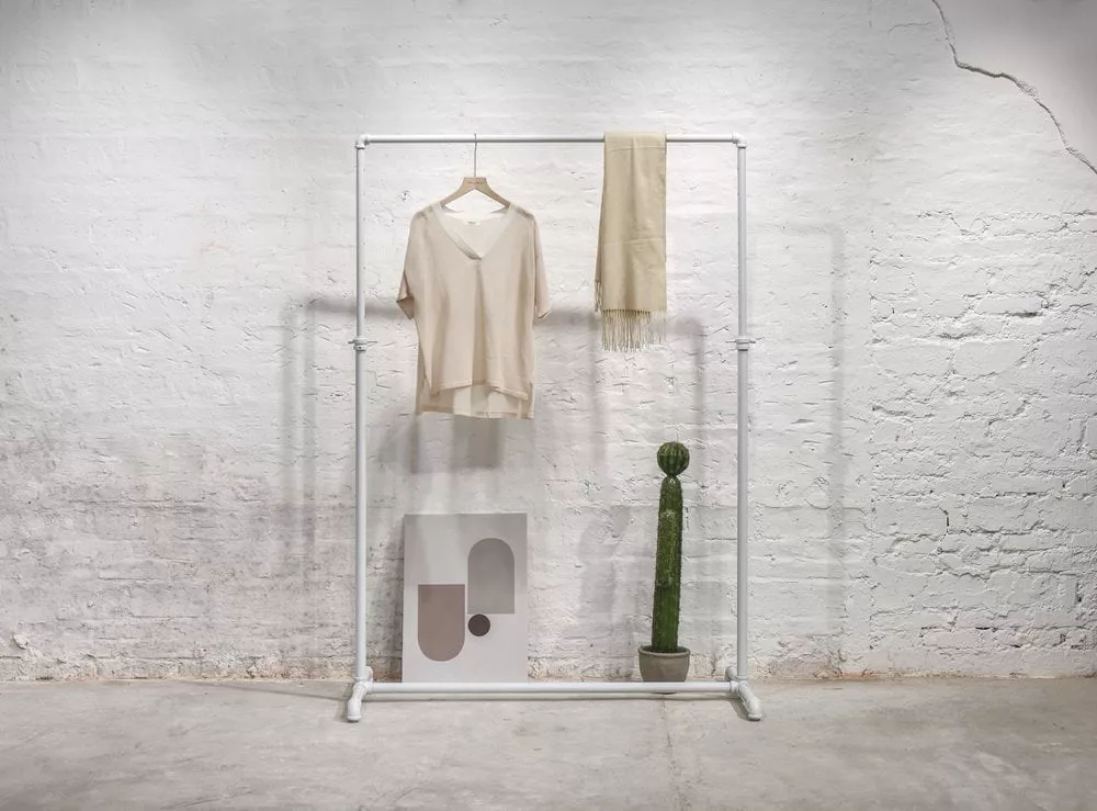 A White Adjustable Pipe Rack is positioned against a white brick wall, gracefully showcasing a beige sweater on a hanger along with a coordinating scarf. Below, an abstract art piece rests against the wall alongside a potted cactus, enhancing the minimalist vibe of the room.