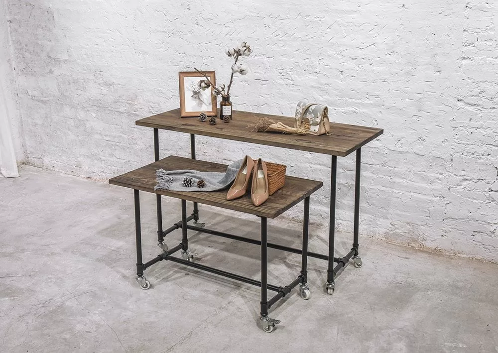 A Small Black Pipe Rack Display Table, featuring two stacked wooden surfaces on wheels, is artfully adorned with a framed photo, cotton plant, bottle, shoes, scarf, and various accessories. The sleek black pipe design enhances the rustic charm of this stylish setup against a textured white brick wall and concrete floor.
