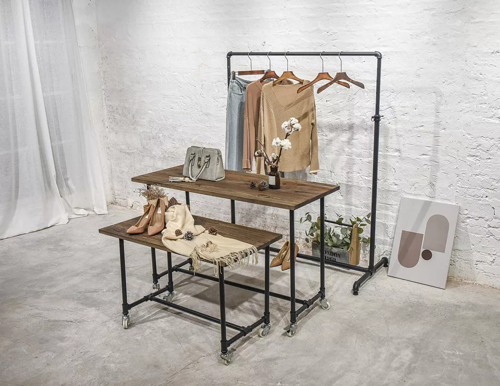 The display in the chic boutique showcases a Small Black Pipe Rack Display Table, elegantly accessorized with beige garments. Nearby, you can find shoes, a handbag, and decorative items such as a vase filled with flowers. The white brick wall and minimalistic art piece provide the perfect backdrop for this stylish setting.