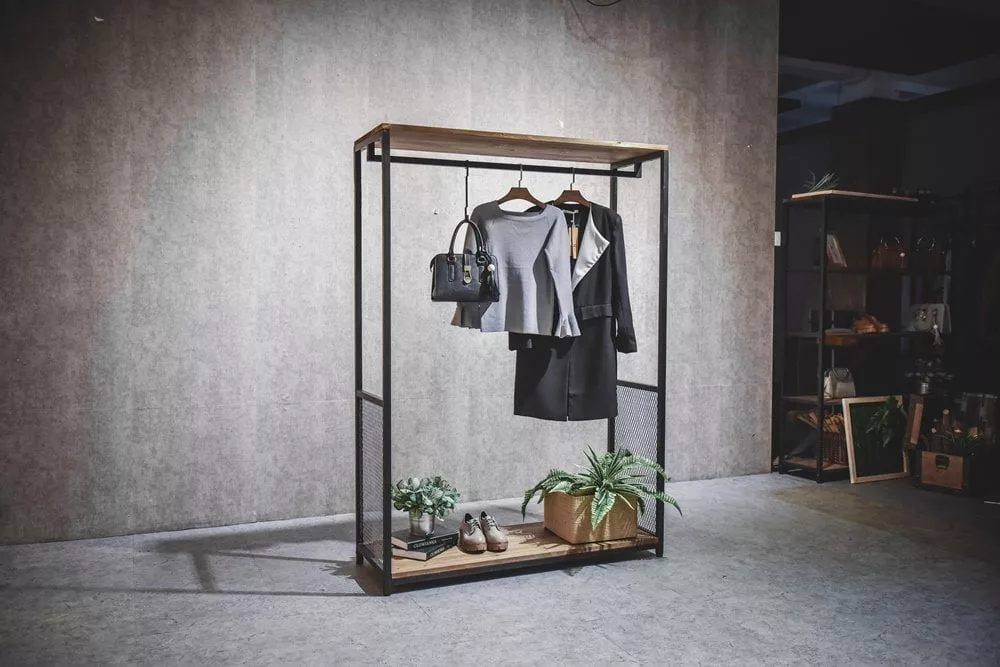 An Industrial Display Rack with a Timber Top and Base stands in a minimalist room. The wooden top and base support a gray top, black skirt, handbag, and coat. The lower shelf holds a plant and shoes, all set against a textured gray wall backdrop.