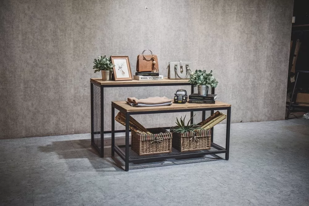 A stylish room features the Industrial Display Table Set, showcasing two stacked wooden tables framed in metal. The top table is adorned with plants, a framed picture, books, and decor, while the lower table holds baskets with rolled towels and small potted plants against a textured gray wall.