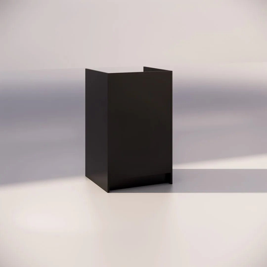 A minimalist, black, rectangular podium or lectern stands on a light gray floor against a plain wall. Strong lighting casts distinct shadows, highlighting the sleek design and simple geometry of the object.
