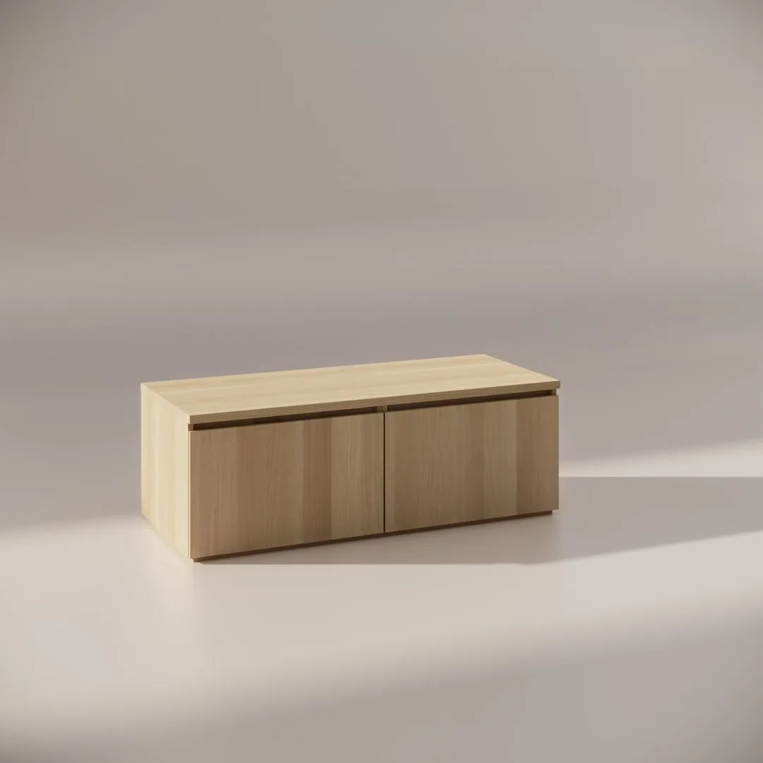 A minimalist wooden storage bench with a rectangular shape and subtle grain texture sits in a softly lit space, casting gentle shadows on the surface. The bench features two panel doors and a smooth top.